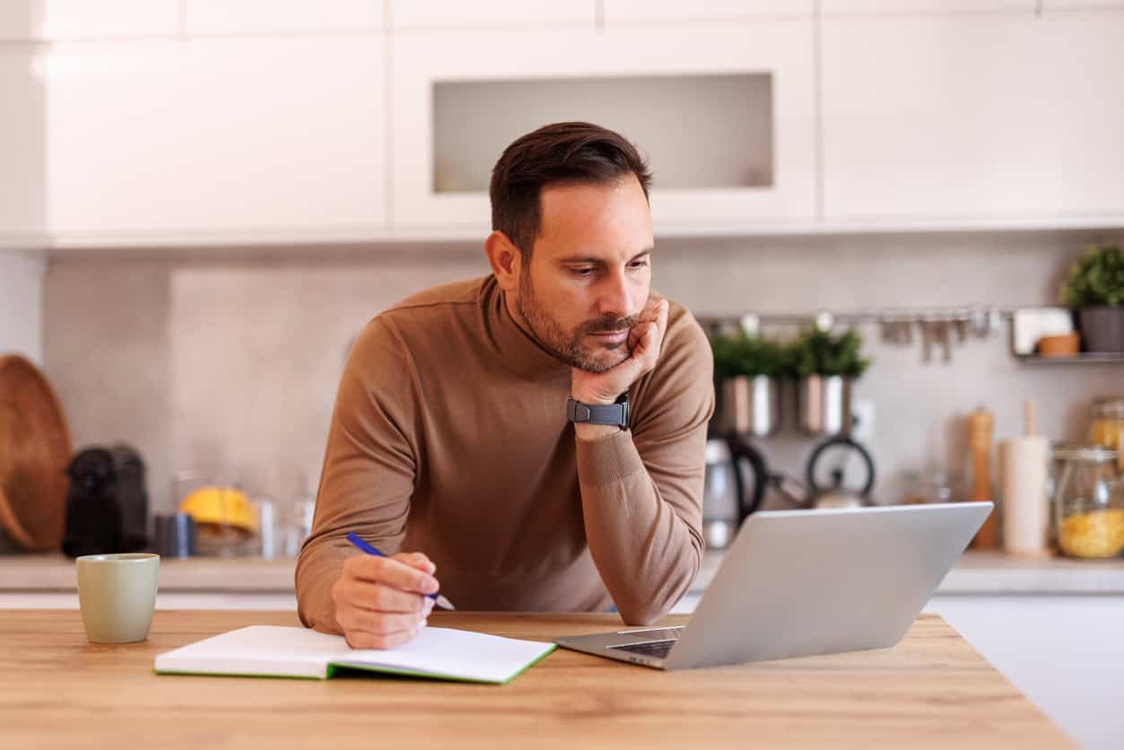 A man reading info on his laptop while writing in note pad at home.